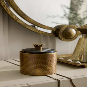 A cylinder of rich hammered brass is juxtaposed with a black marble lid accented with a sculpted brass handle inspired by the textural shape of a pomegranate calyx.