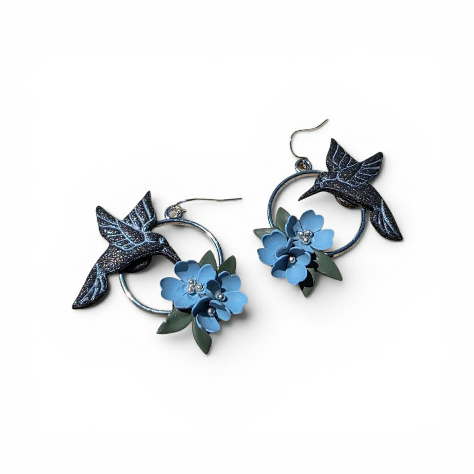 Handcrafted polymer clay hoop earrings featuring sculpted hummingbirds, delicate blue blooms, and leafy details. Lightweight, dimensional, and inspired by the beauty of a garden in motion.